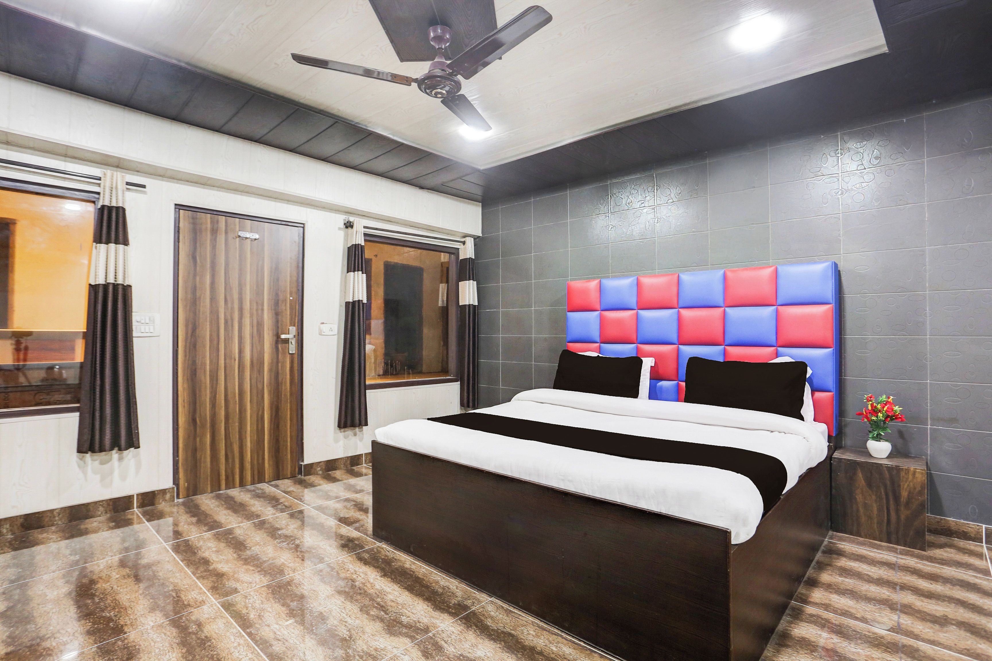 Habitación de hotel con una cama, ventilador de techo, ventana con cortinas y piso de baldosas.