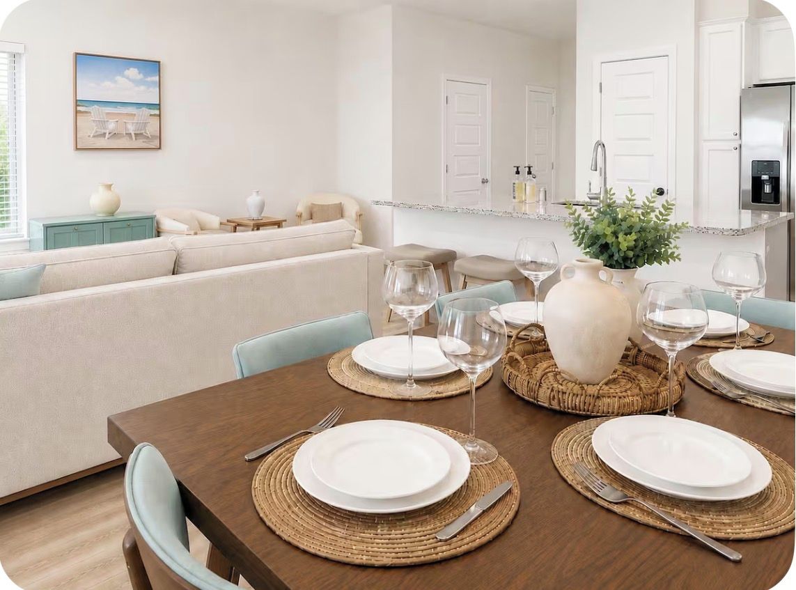 Un comedor con una mesa de madera dispuesta para cuatro personas, rodeada de sillas, y una cocina al fondo.