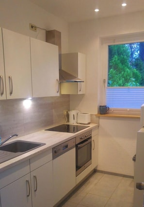 Cocina compacta con armarios blancos, horno empotrado, microondas y ventana con vistas a zonas verdes.
