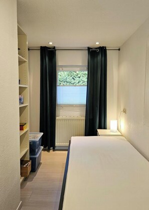 Un dormitorio con cama, una mesita de noche, una estantería con libros, una ventana con cortinas y un radiador.