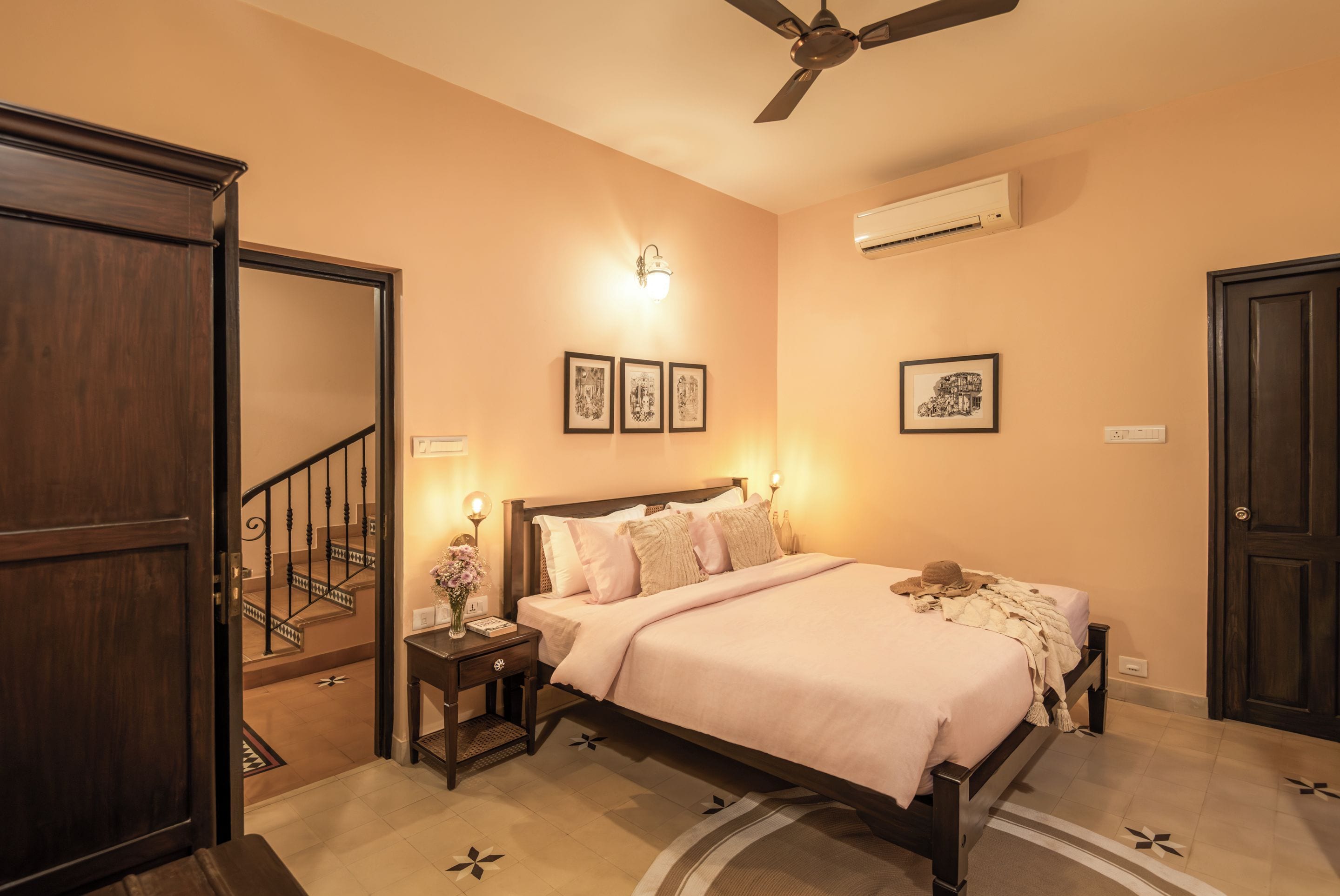 A bedroom with a bed, nightstand, wall-mounted lights, framed pictures, a ceiling fan, and an air conditioning unit.