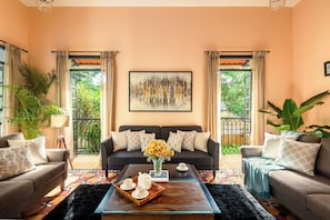 A living room with a sofa set, a coffee table with a tray of cups, a painting on the wall, and a view through the windows. - Galeria  Portuguese style 4bhk Pvt Pool (Arpora)