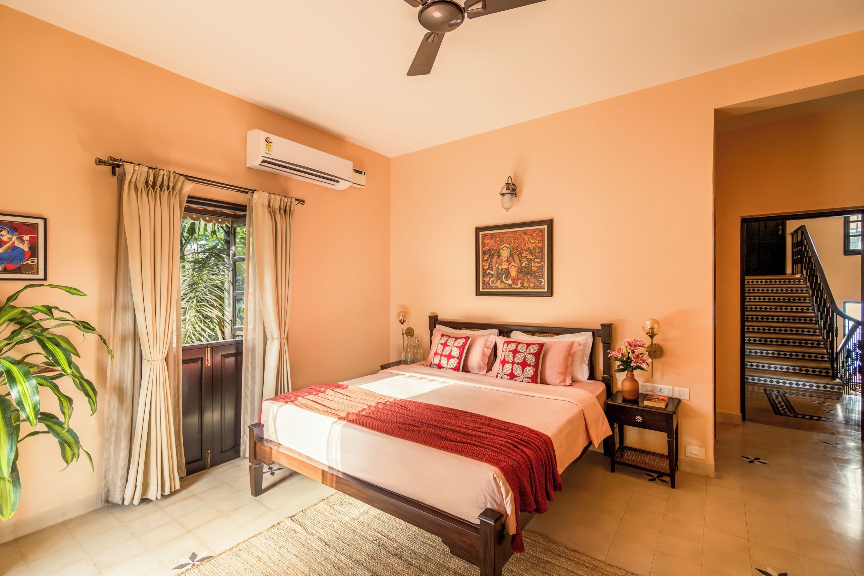 Habitación de hotel con cama, mesita de noche, aire acondicionado, ventana con cortinas, un cuadro, una escalera y una planta en maceta.