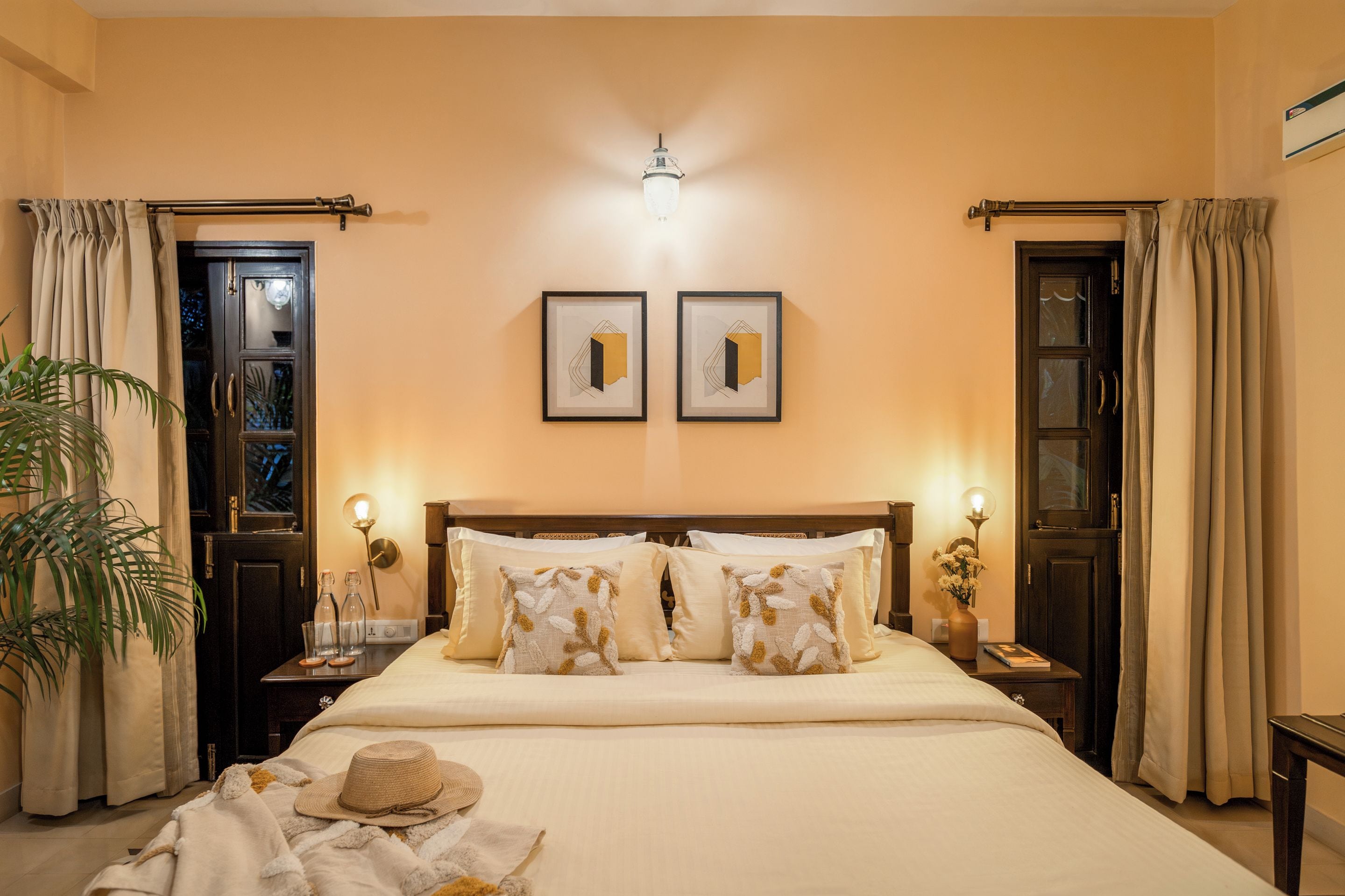 A hotel room with a large bed, two bedside tables, a nightstand, a potted plant, and two framed pictures on the wall.