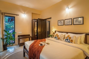 A hotel room with a bed, a bedside table, a chair, a potted plant, and a door leading to a balcony. - Galeria  Portuguese style 4bhk Pvt Pool (Arpora)