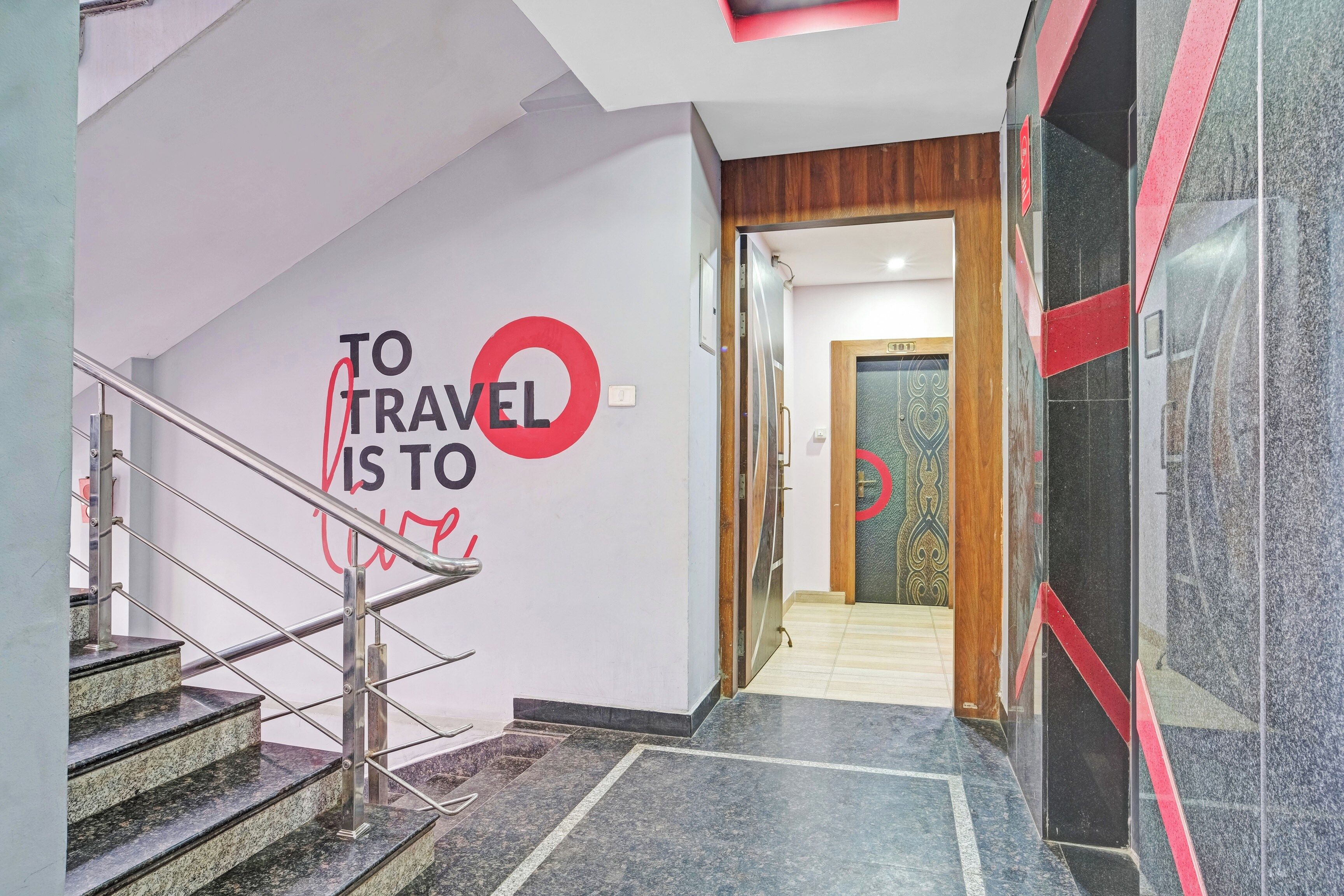 A hotel lobby with a staircase, a wall with a travel-themed sign, and a wooden door leading to another room.