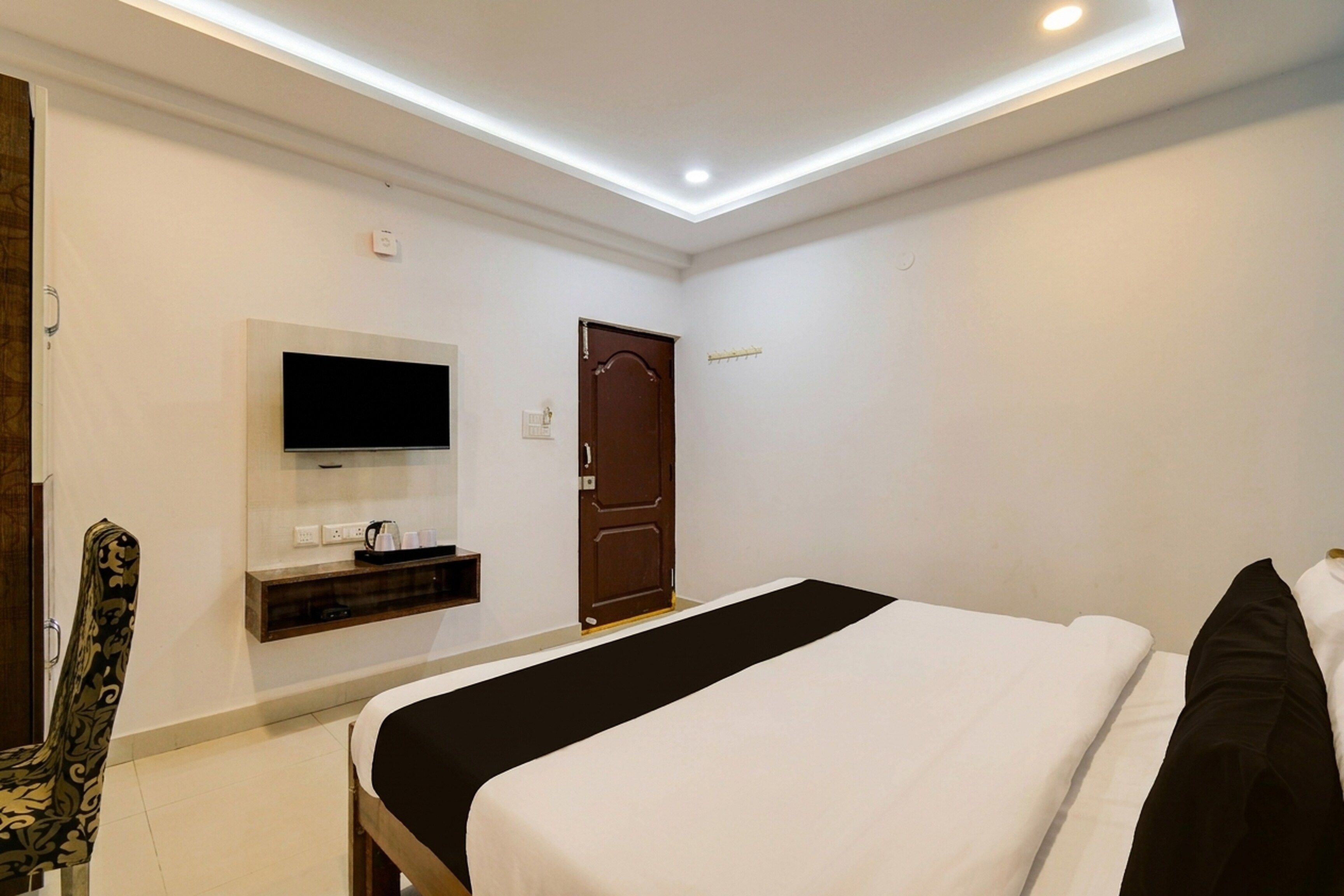 Habitación de hotel con una cama, televisor montado en la pared, una puerta y un estante pequeño con objetos.
