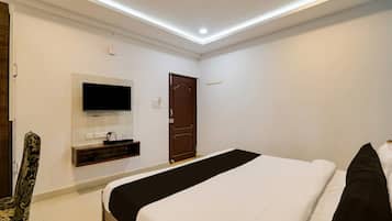 Habitación de hotel con una cama, televisor montado en la pared, una puerta y un estante pequeño con objetos.