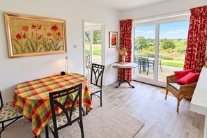 A room with a dining table, chairs, a small table with a vase, and a wicker chair with red cushions. - Willow Bank, Low Row, bungalow with stunning countryside views to Hadrian's Wall (Low Row)