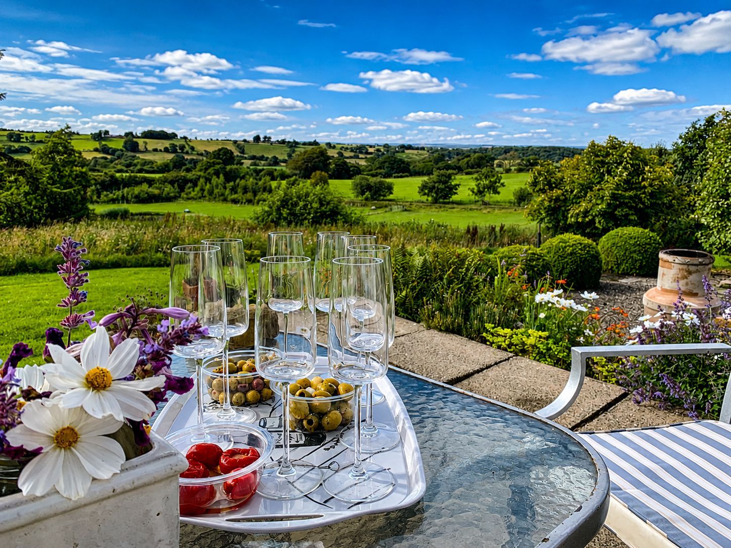 A table set outdoors with wine glasses and olives, surrounded by flowers and greenery.