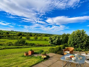 An outdoor dining area with a table set for two, overlooking a lush green landscape and rolling hills under a partly cloudy sky. - Willow Bank, Low Row, bungalow with stunning countryside views to Hadrian's Wall (Low Row)