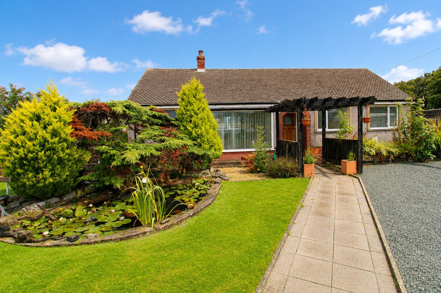 Una casa con un jardín bien cuidado, un estanque con nenúfares y un cielo azul despejado.