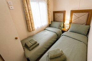 A room with two beds, a nightstand with a lamp, and a window with curtains. - Calf Lonning Caravan, Plumbland, in a quiet Cumbrian village close to Keswick and Western Lakes (Plumbland)