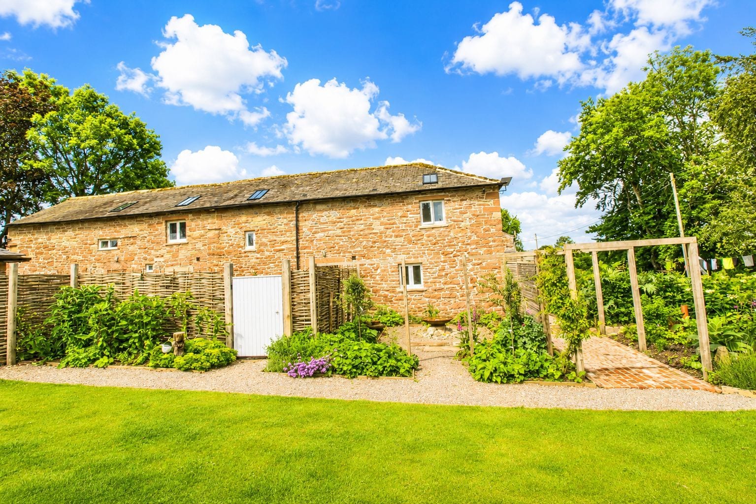 A stone house with a white door, a garden with various plants, and a clear blue sky.
