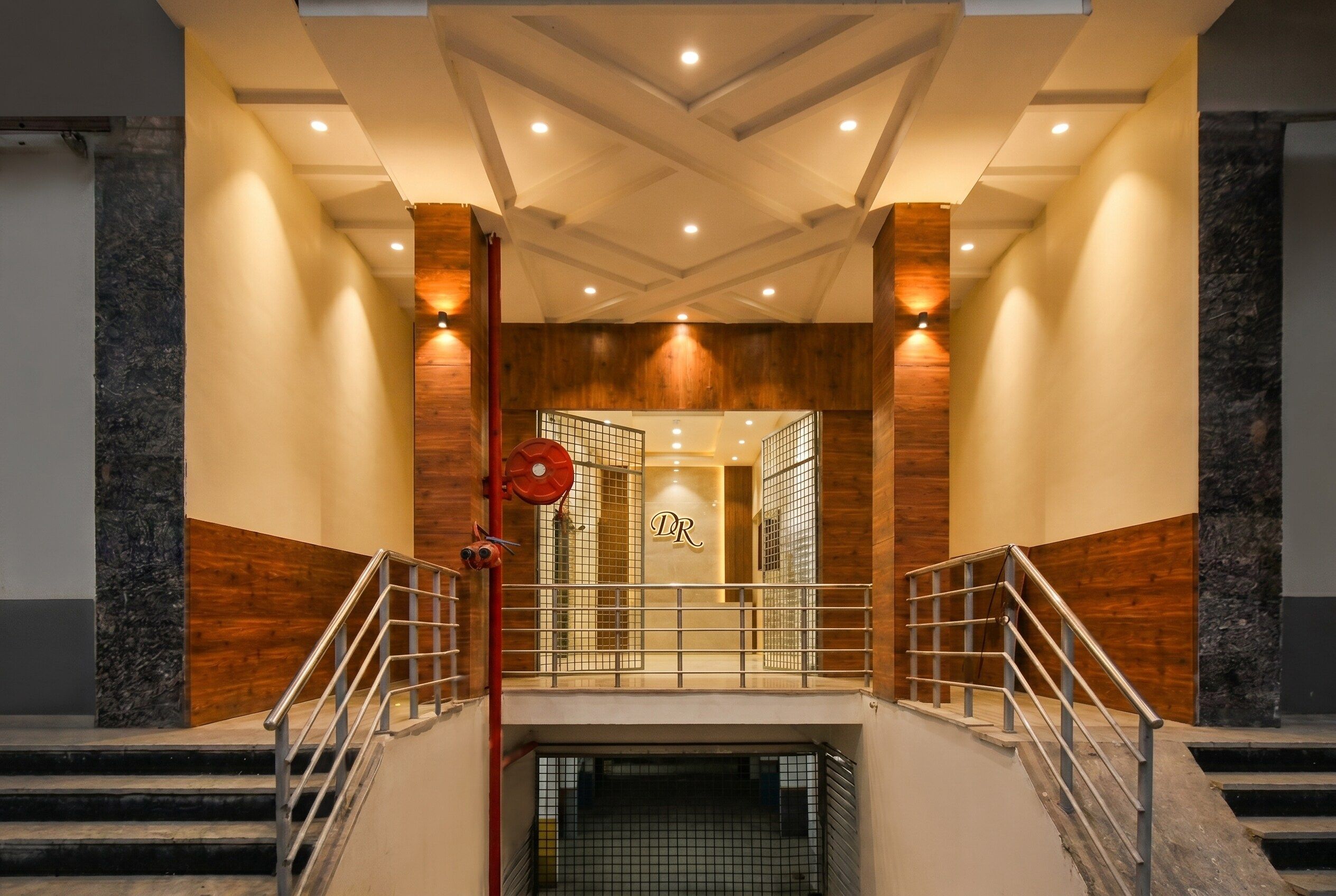 Entrada de un hotel con diseño moderno, que presenta una escalera central con barandas de metal, una pared de madera e iluminación empotrada en el techo.