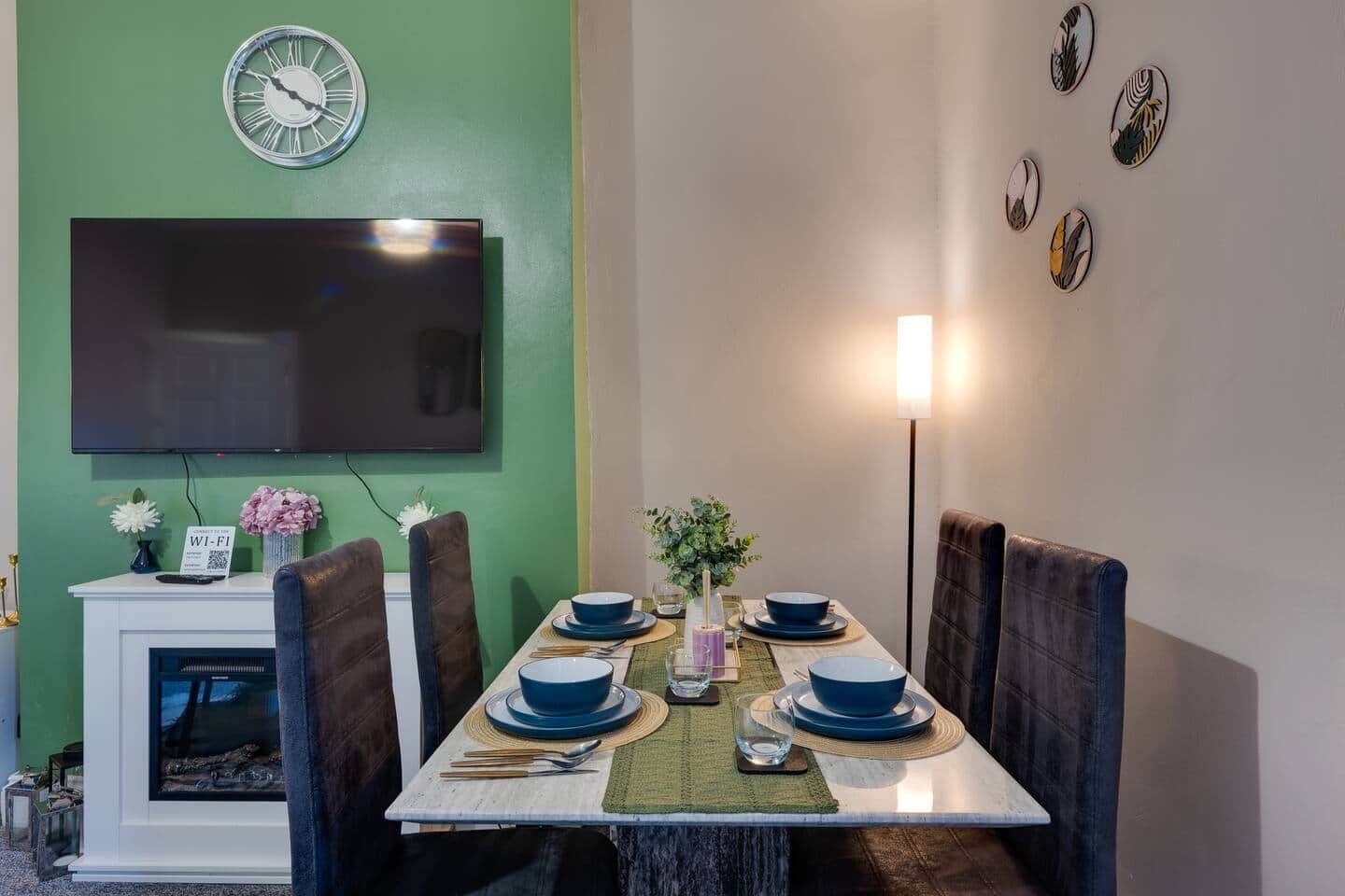 A dining area with a table set for four, a flat-screen TV mounted on a green wall, and a fireplace.