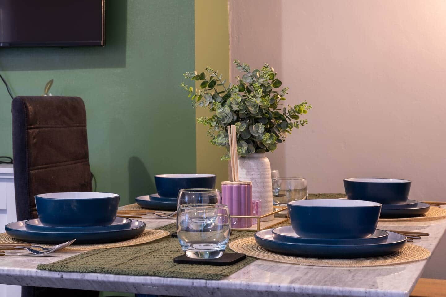 A dining table set with blue plates, bowls, and cutlery, accompanied by a vase with green foliage.