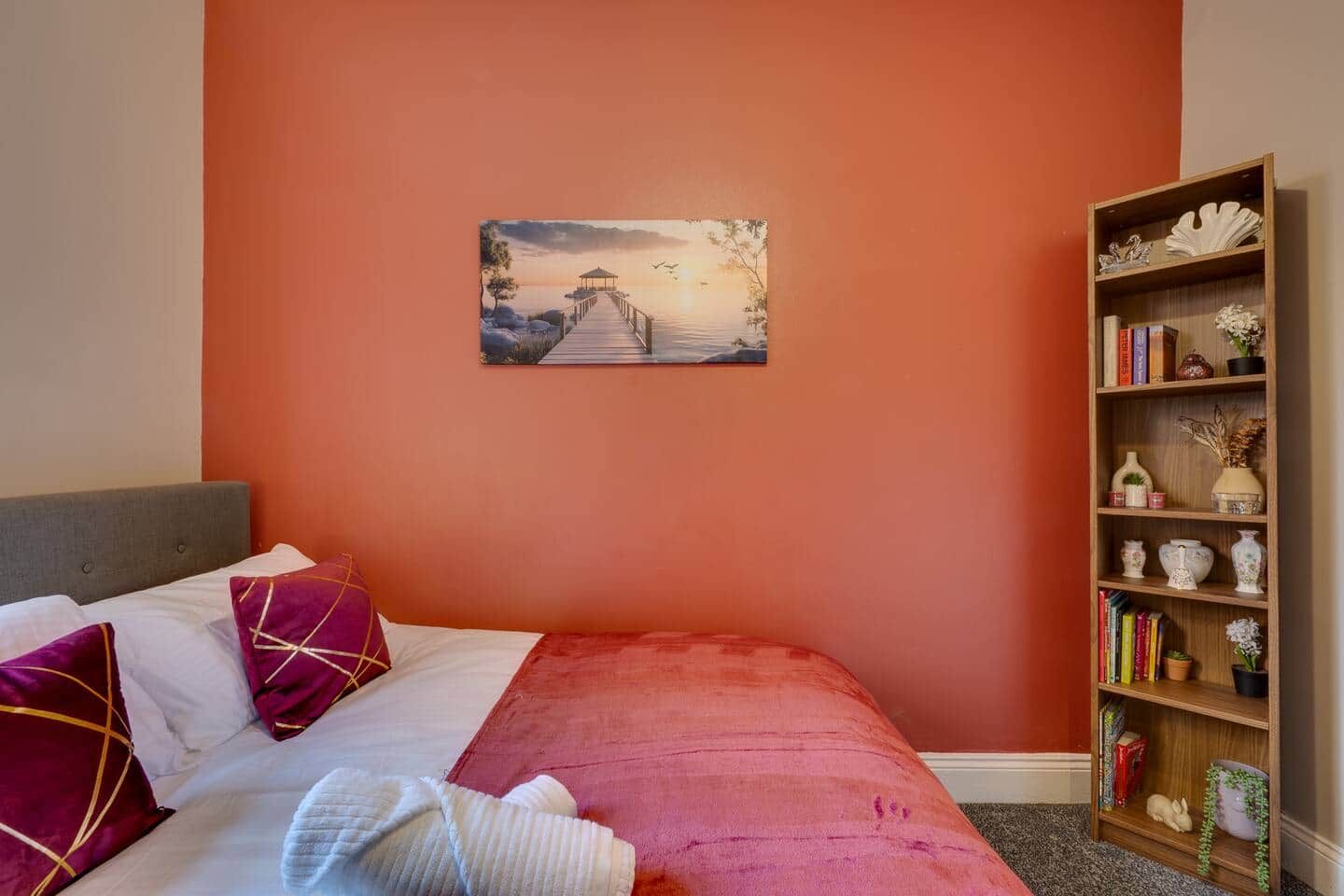 Een slaapkamer met een bed, kussens, een schilderij en een boekenkast.