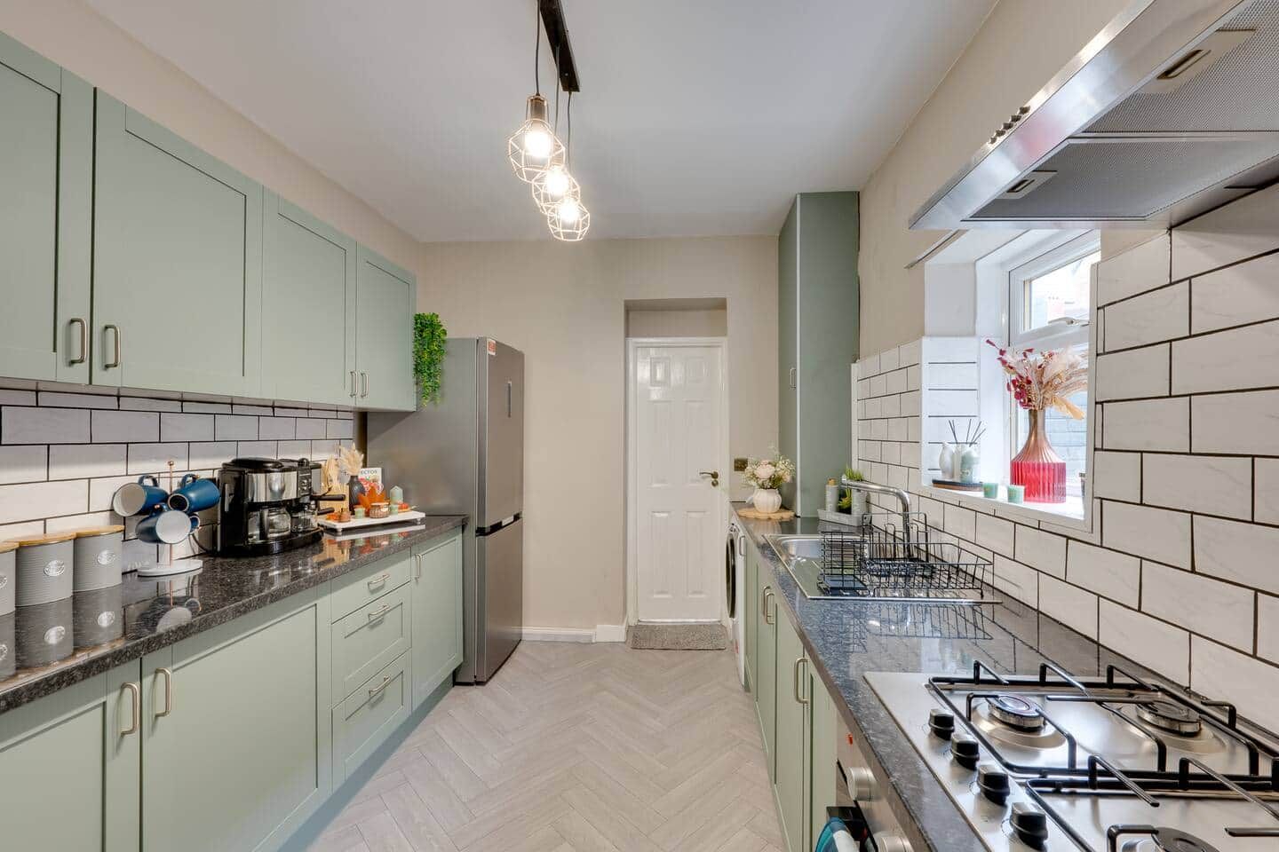 A modern kitchen with light green cabinets, a stainless steel refrigerator, a gas stove, and a window with a view.