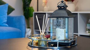 Um candeeiro de vidro com a inscrição 'HOME Sweet HOME', colocado sobre uma mesa, com um sofá azul e uma planta ao fundo.
