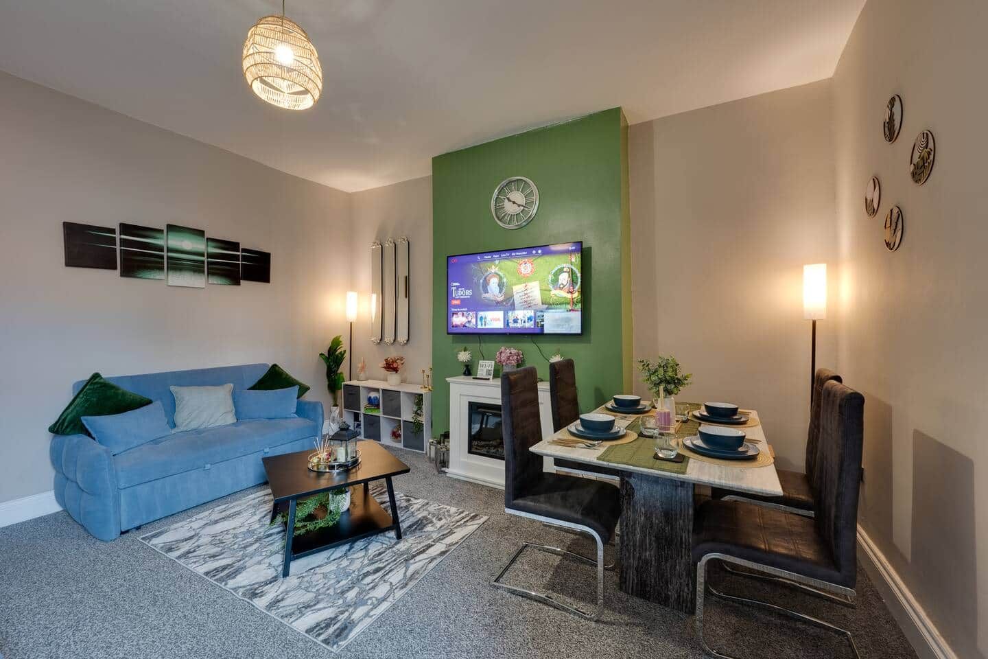 Een moderne woonkamer met een blauwe bank, een eettafel gedekt voor vier personen, een open haard en een flatscreen televisie aan de muur.