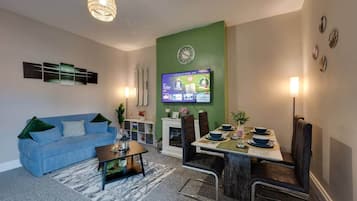 Een moderne woonkamer met een blauwe bank, een eettafel gedekt voor vier personen, een open haard en een flatscreen televisie aan de muur.