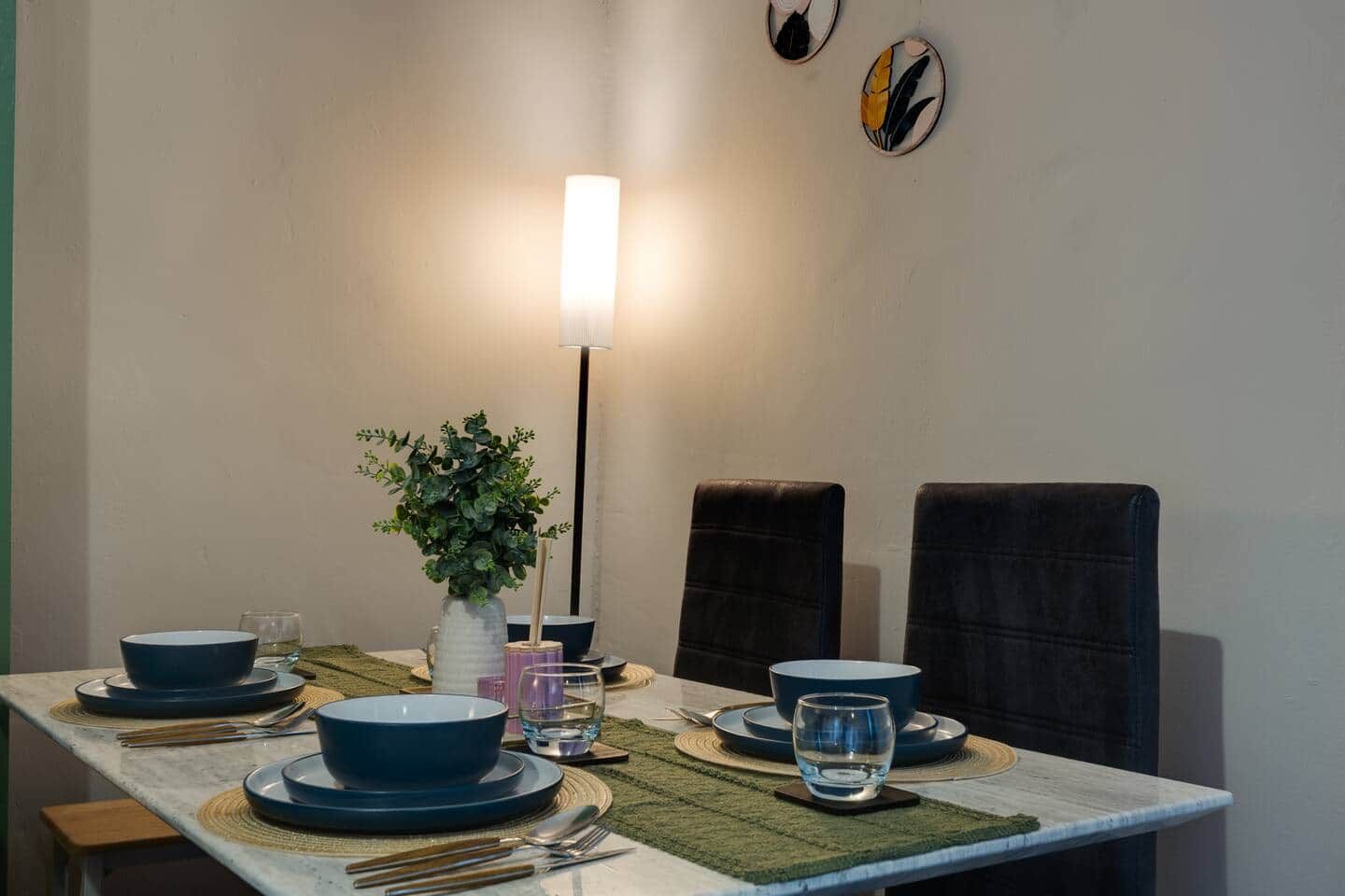 Een eettafel gedekt met borden, kommen, glazen en bestek, met een vaas groene planten en een hoge vloerlamp.