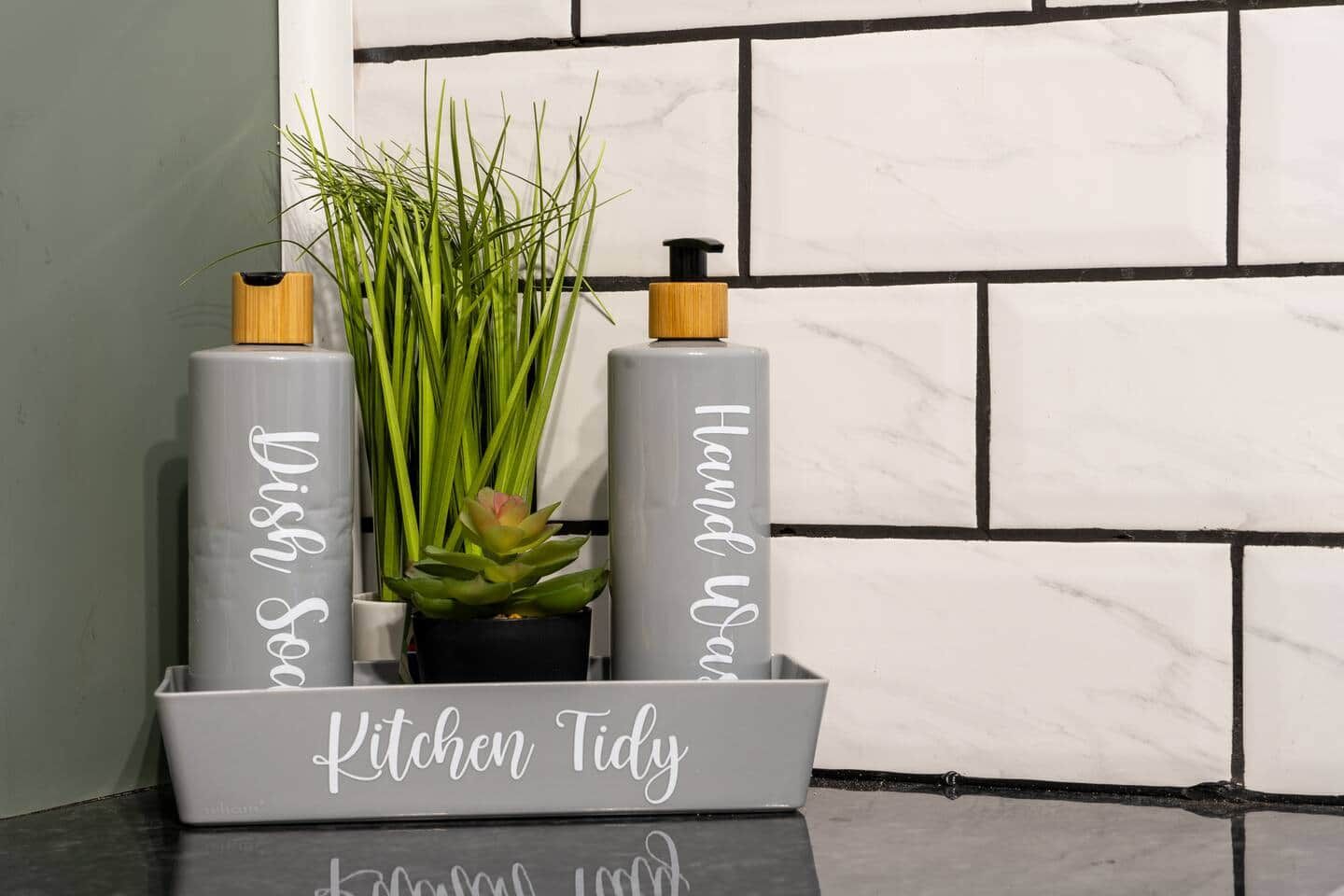 A kitchen area with a tiled backsplash, a grey kitchen trolley with the text 'Kitchen Tidy,' and two cylindrical containers with pumps.