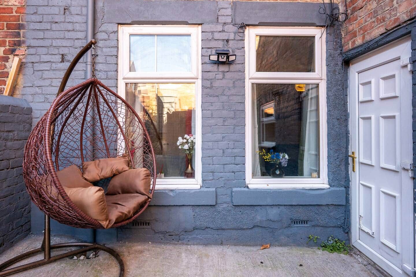 A hanging chair in a courtyard with a brick wall, two windows, and a door.