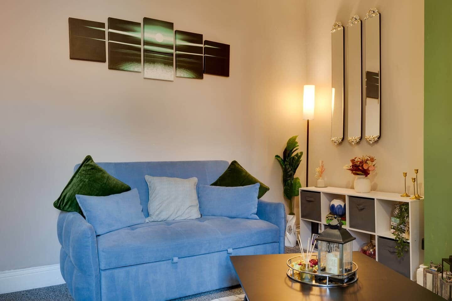 A living room with a blue sofa, a coffee table with a tray, and a wall with three framed artworks and a mirror.