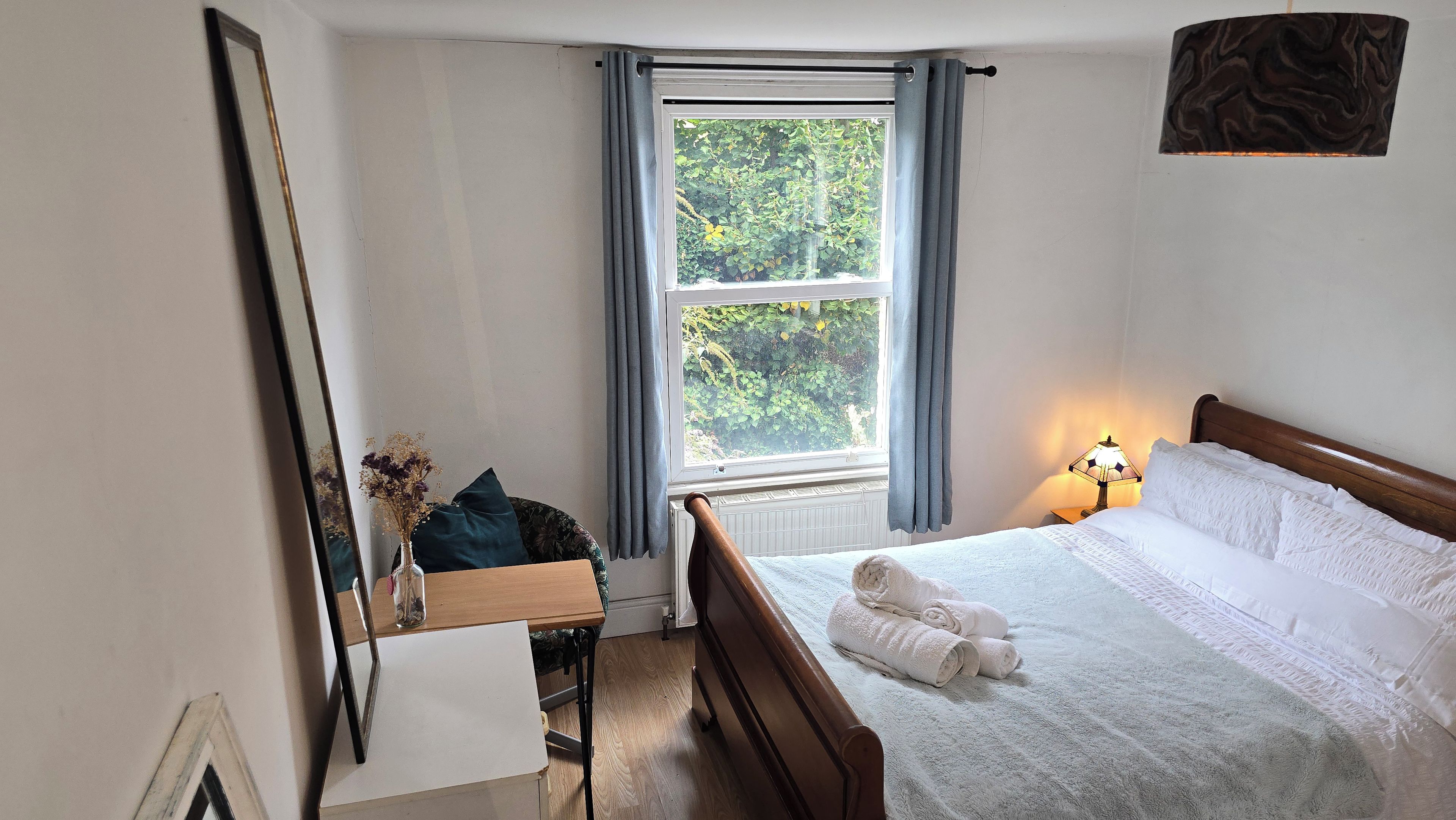 A bedroom with a bed, a bedside table with a lamp, a window with curtains, and a mirror on the wall.