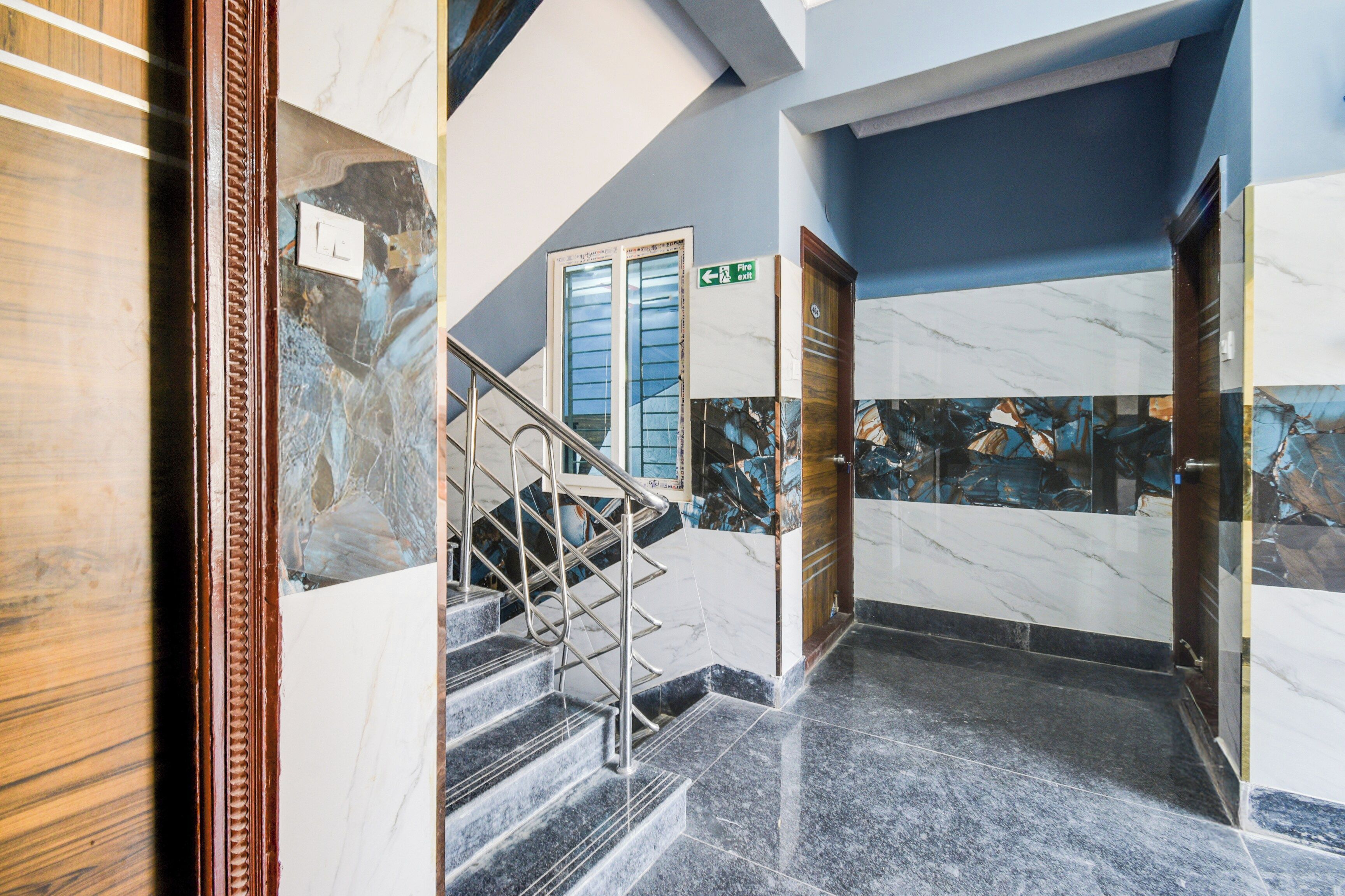 A marble-floored hallway with a staircase, a fire exit sign, and a door with a wooden frame.