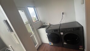 A compact laundry room with a front-loading washer and dryer, a small sink, and a window providing natural light. - Pukekohe 3 Palms 45 Hooper Ave (Pukekohe)
