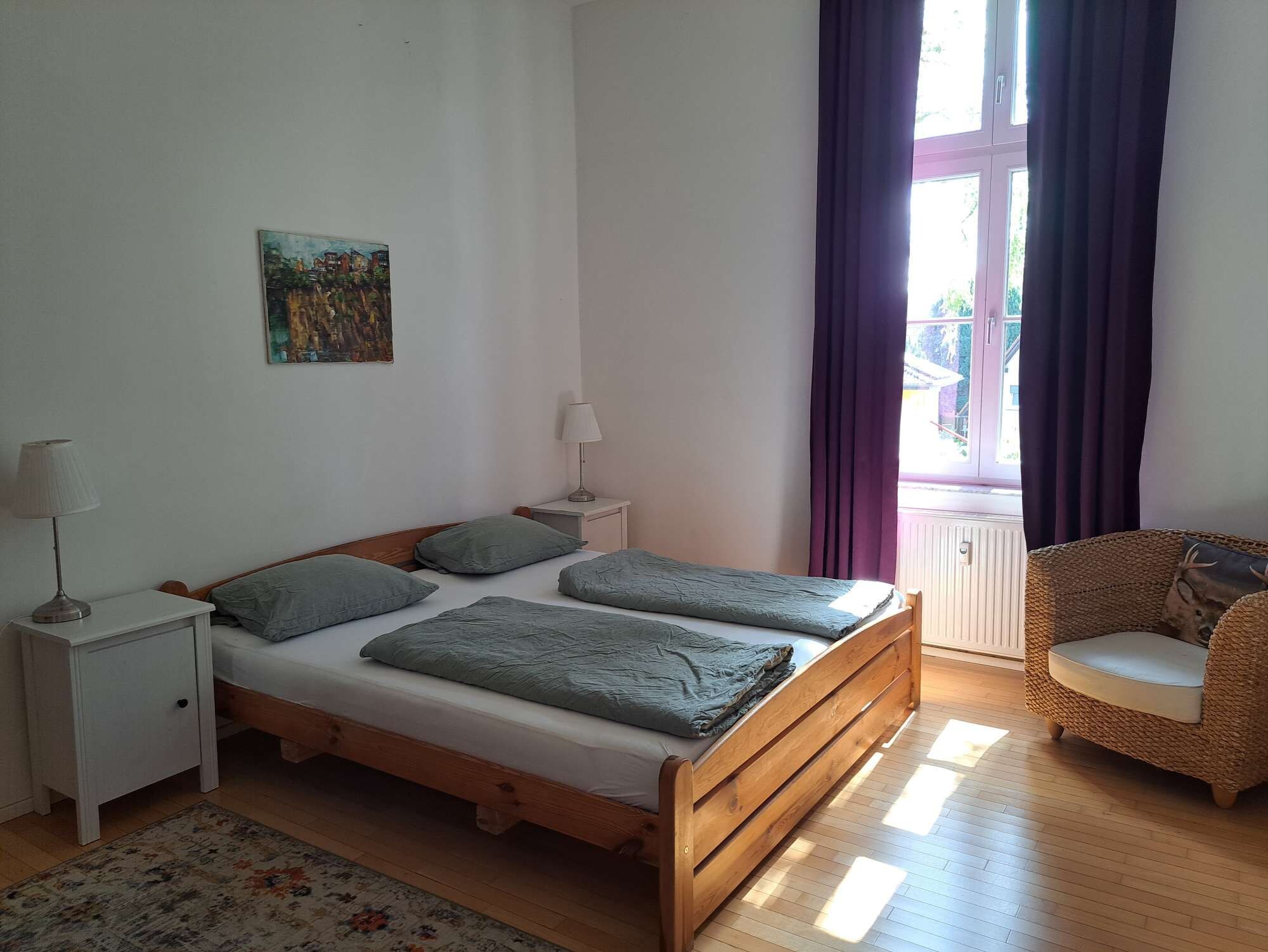 Ein Schlafzimmer mit einem hölzernen Bett, einem Nachttisch, einer Lampe, einem Bild an der Wand und Blick durch ein Fenster mit Vorhängen.