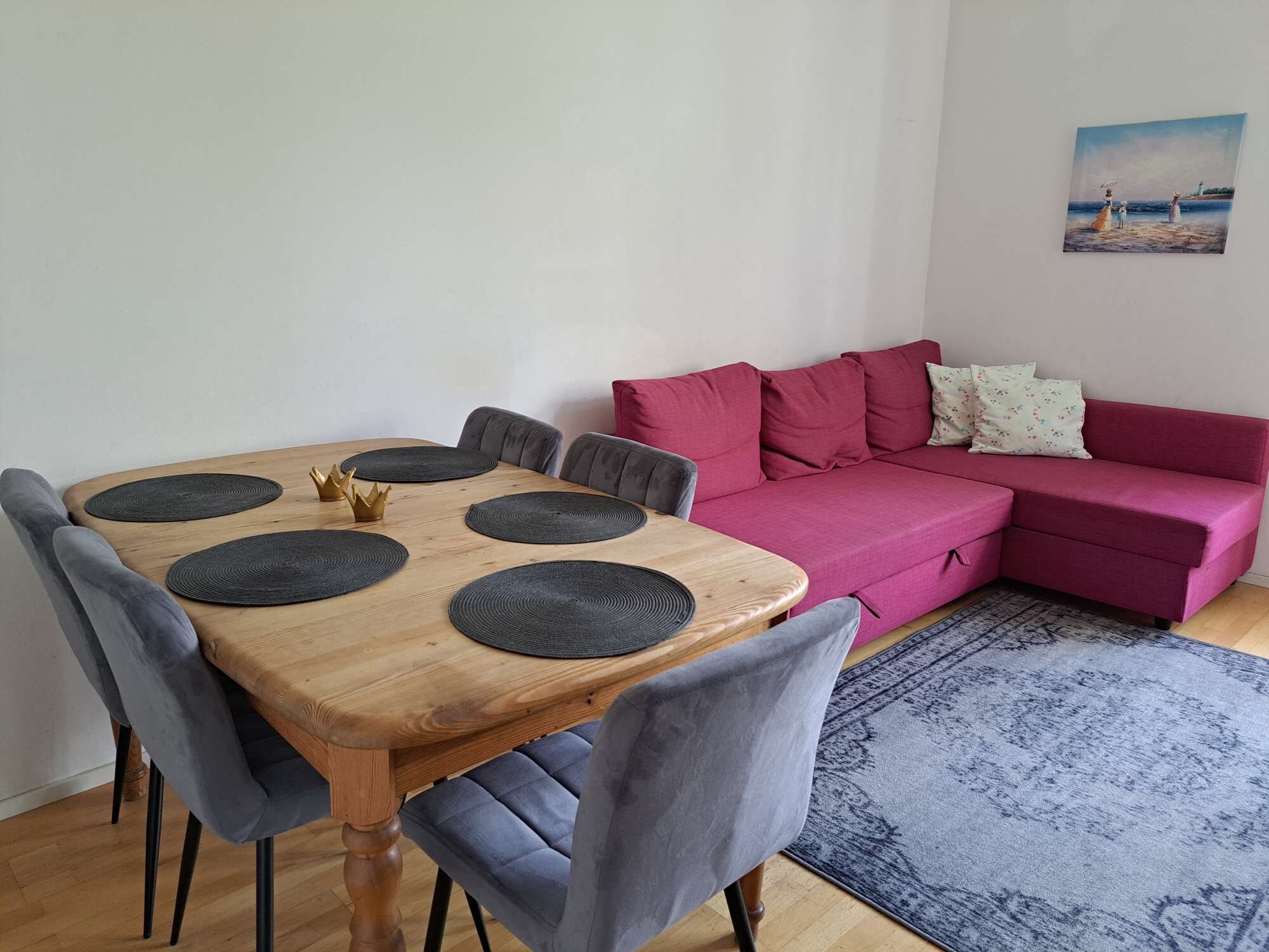 Ein Zimmer mit einem Holztisch, sechs grauen Stühlen, einem rosa Sofa und einem blauen Teppich mit Muster.