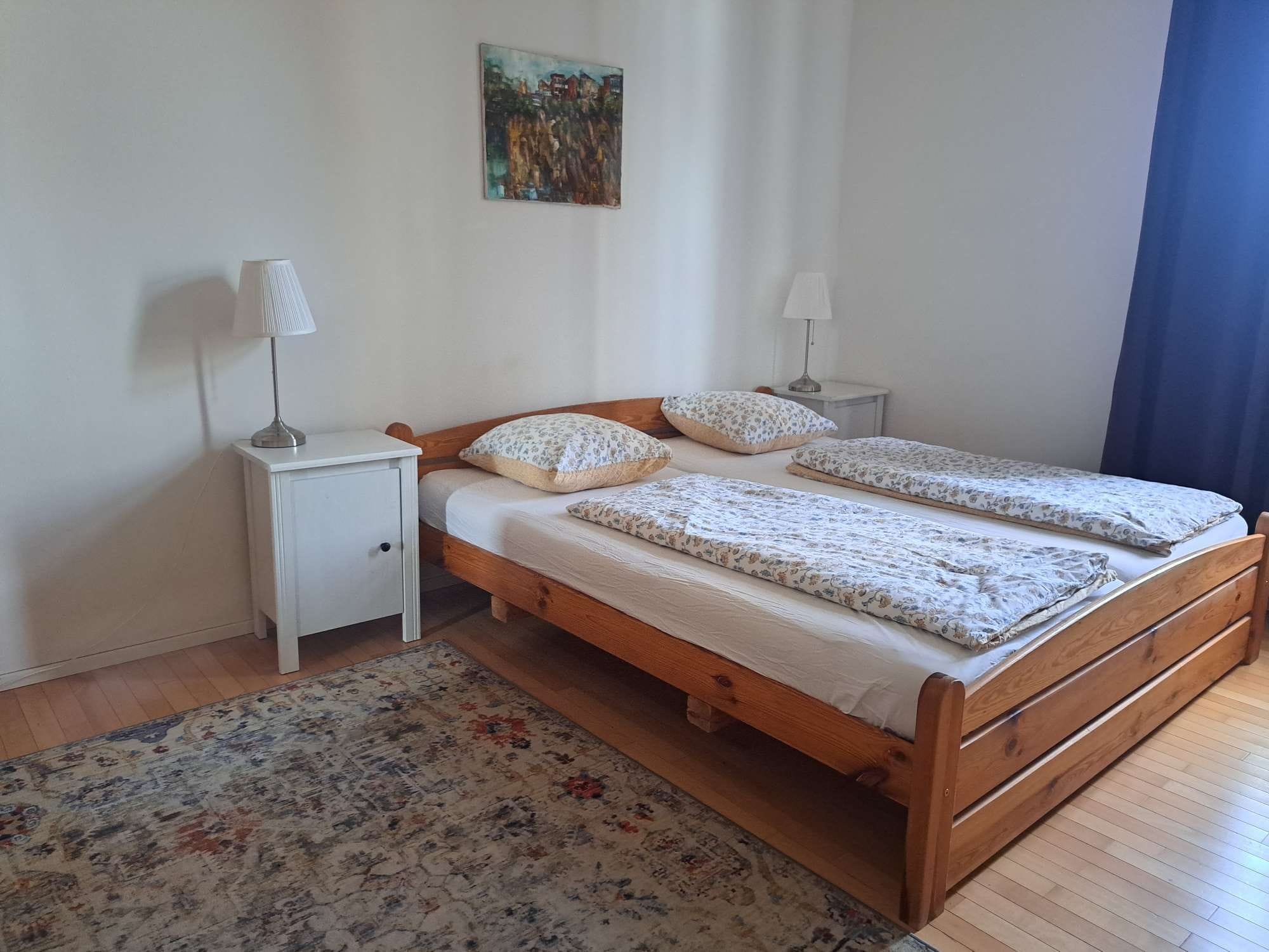 Ein Schlafzimmer mit einem hölzernen Bett, zwei Nachttischen, einem kleinen Tischchen, einem blumigen Teppich und einem Bild an der Wand.