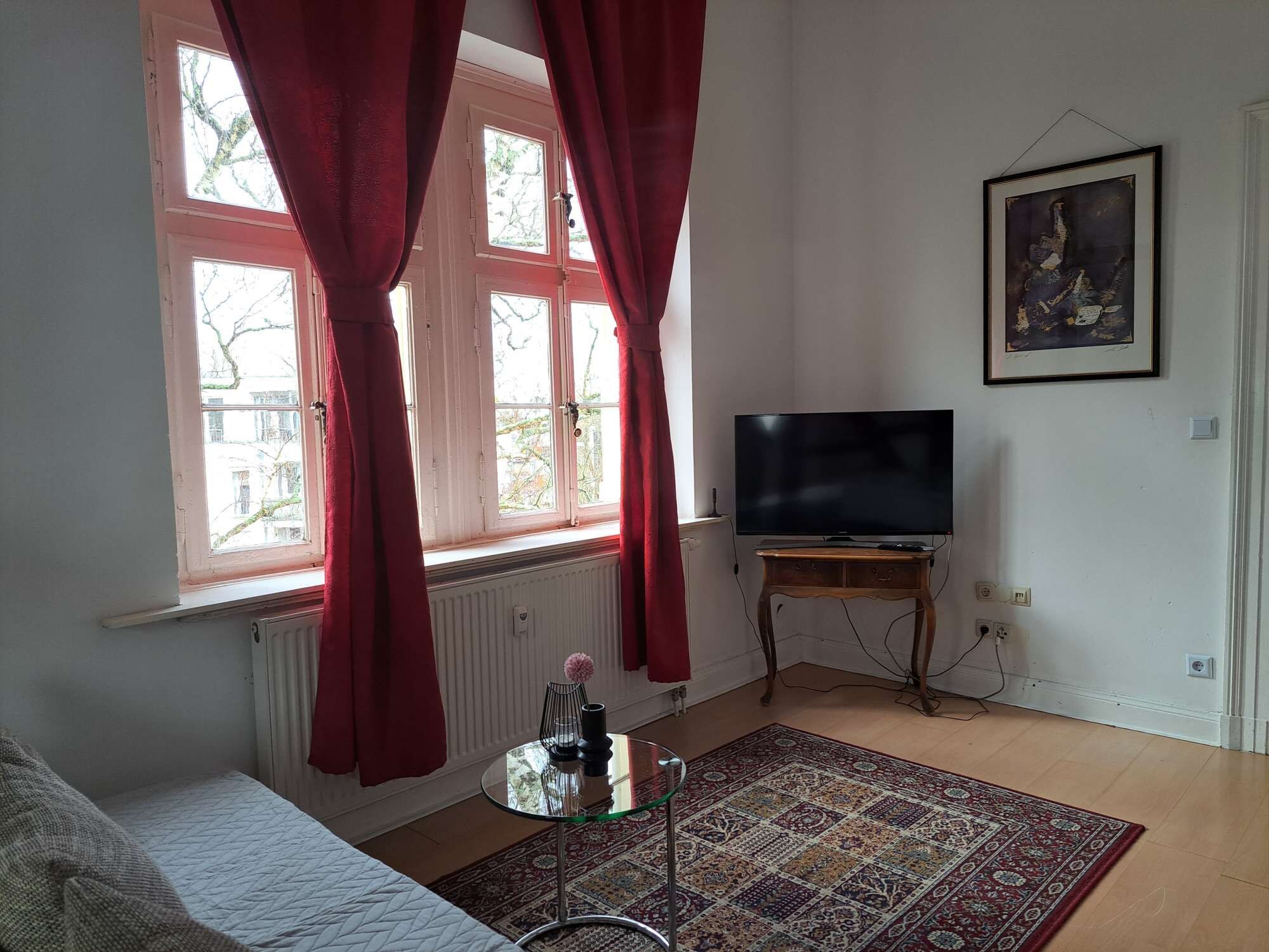 Ein Wohnzimmer mit einer Couch, einem Fernseher auf einem Holzboden, einem gemusterten Teppich und einem Fenster mit Vorhängen.