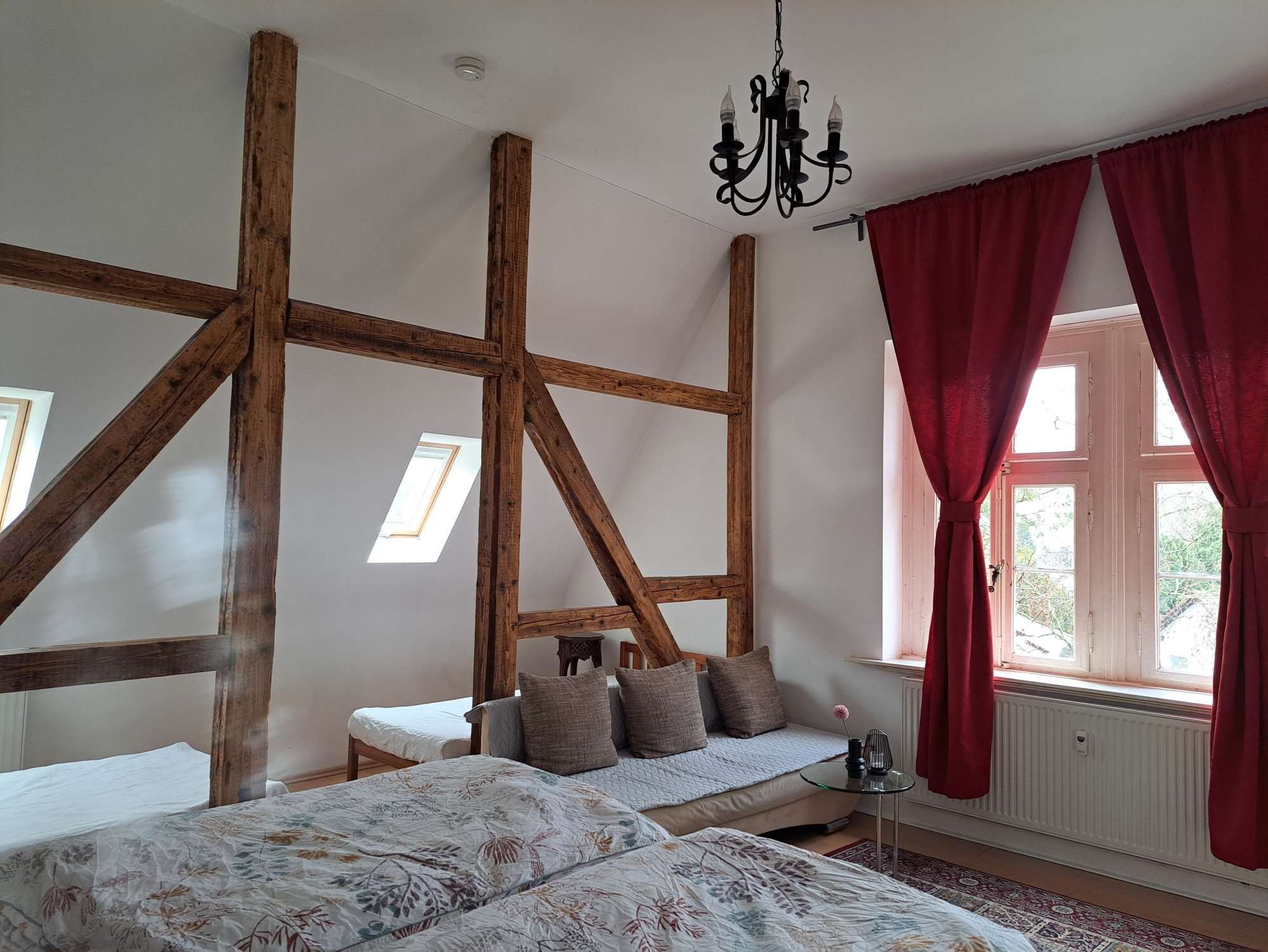 Ein Zimmer mit freiliegenden Holzbalken, einem Bett mit floralem Bettzeug, einem kleinen Tisch und einem Fenster mit roten Vorhängen.