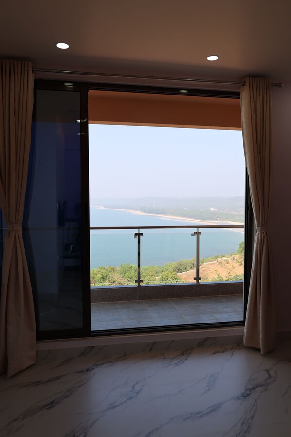 A room with a large sliding glass door leading to a balcony with a view of the sea and distant land.