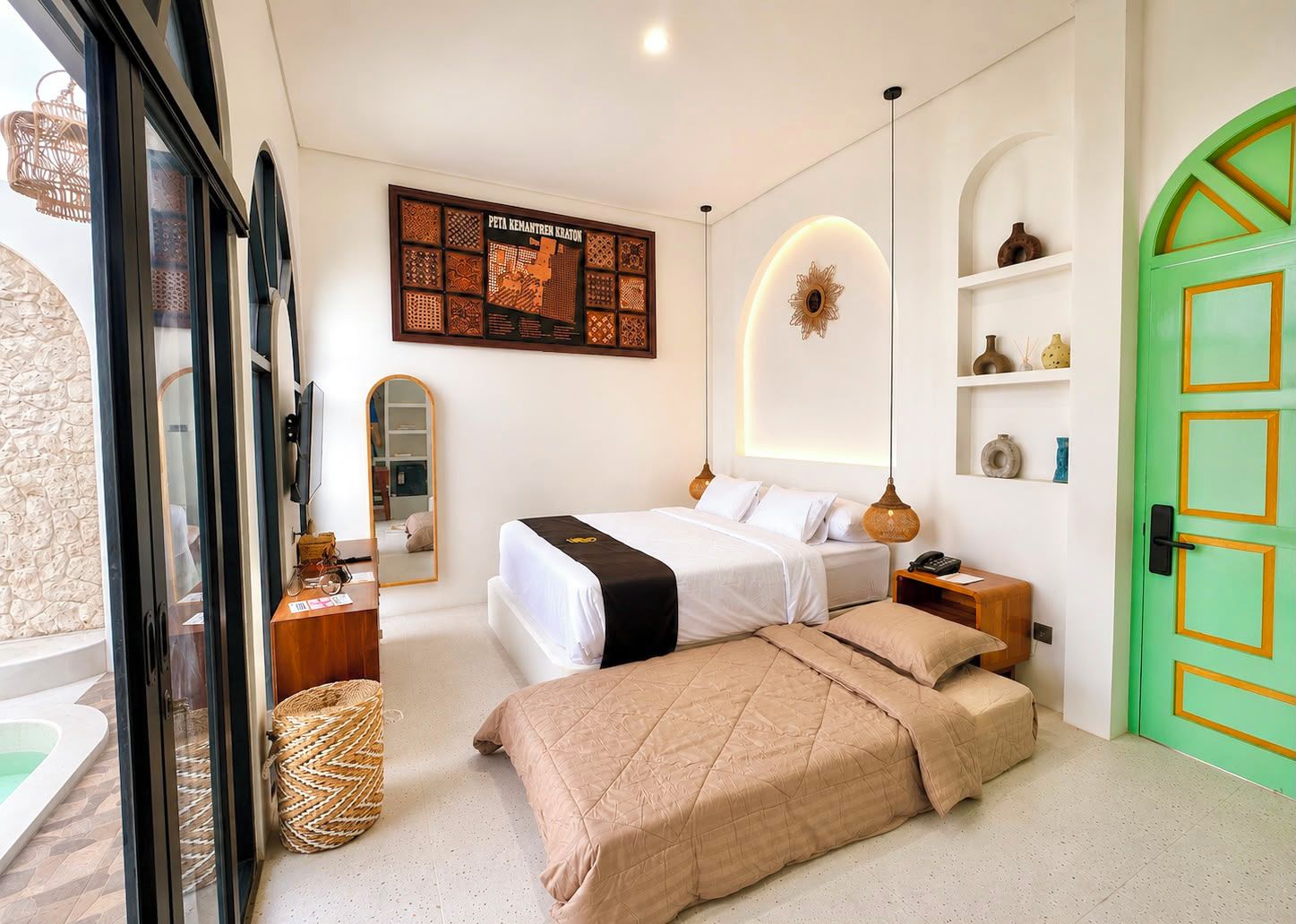 A hotel room with a bed, bedside tables, a mirror, a decorative wall piece, a green door, and a view of the outside.