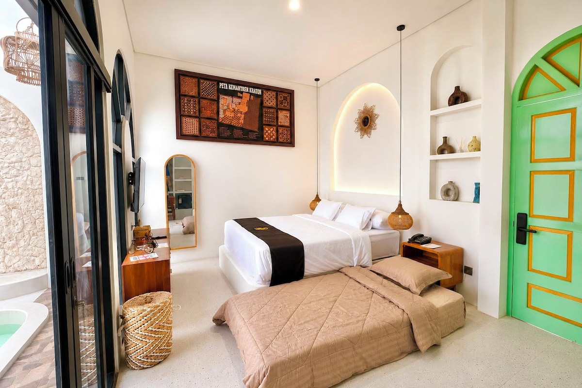 Ein Hotelzimmer mit Bett, Nachttischen, einem Spiegel, einer Wanddekoration, einer grünen Tür und Blick ins Freie.