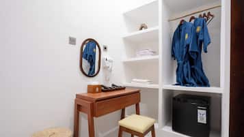 Ein Hotelzimmer mit einem Holztisch, einem Stuhl, einem Spiegel, einem Telefon, einem Regal mit gefalteten Handtüchern, einem Kleiderständer mit Bademänteln und einer schwarzen Mülltonne.