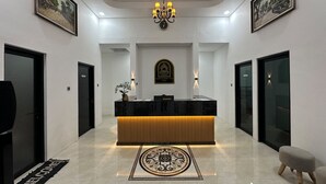 A hotel reception area with a black reception desk, a chandelier, framed artwork on the walls, and a welcome mat. - Mataram Royal Villa Jogja (Kota Yogyakarta)