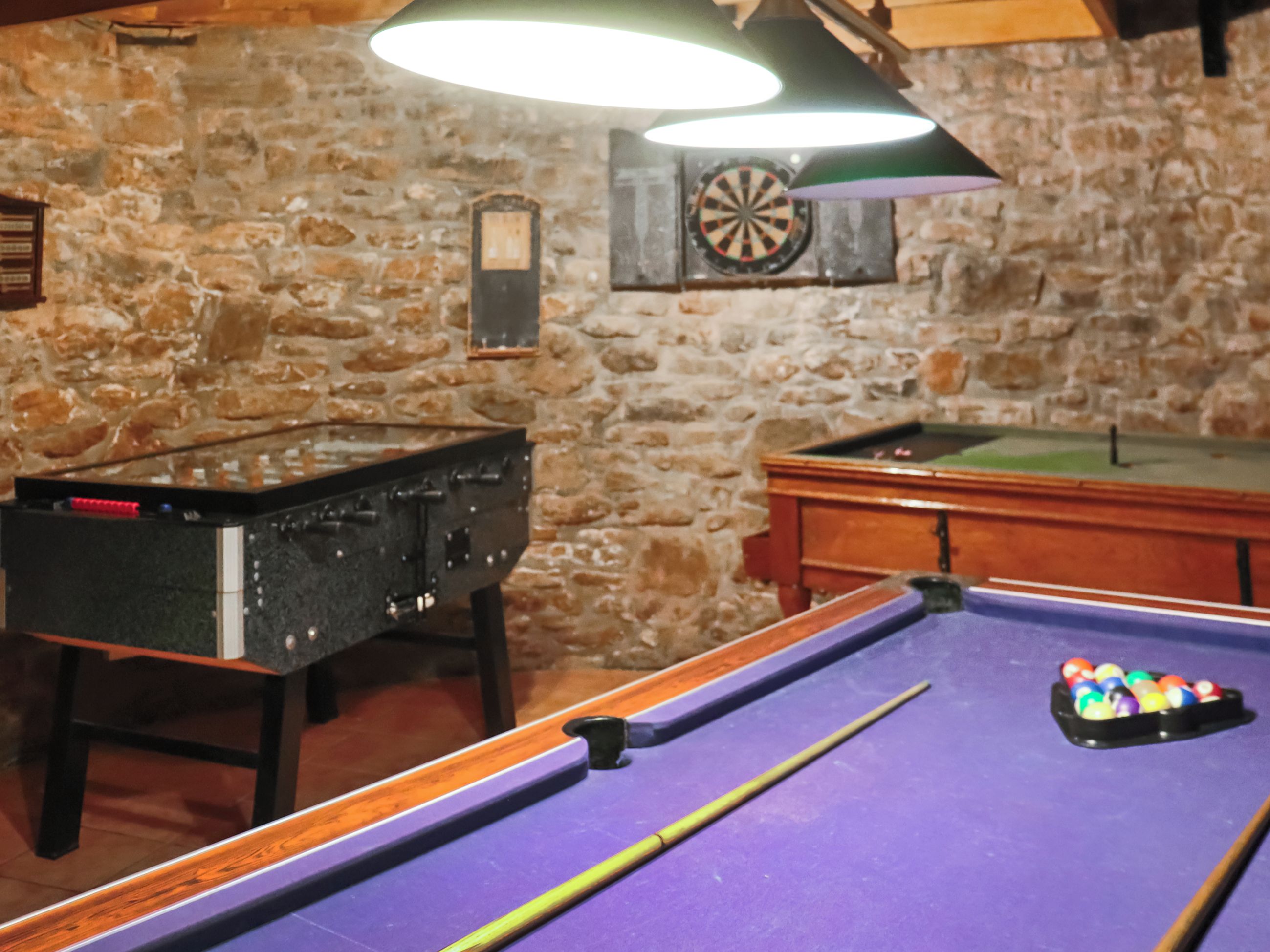 A billiards table with a cue stick, a pool table, and a dartboard in a room with stone walls.