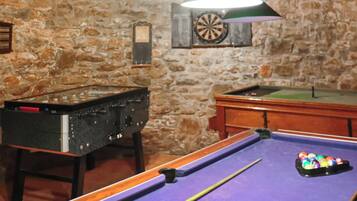 A billiards table with a cue stick, a pool table, and a dartboard in a room with stone walls.