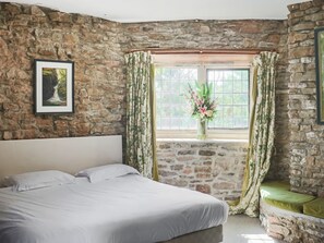 Un dormitorio con pared de piedra, una cama con ropa de cama blanca, una ventana con cortinas florales y un cuadro en la pared.