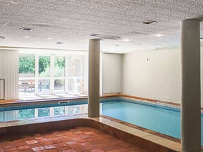 An indoor swimming pool with a tiled floor, large windows, and a view of greenery outside.