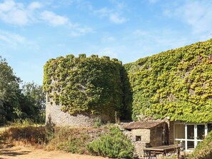 Un edificio de piedra con tejado de tejas, rodeado de vegetación y un cielo despejado.