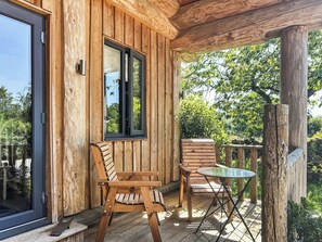 Eine Holzterrasse mit zwei Holzstühlen und einem kleinen Tisch, einer Glastür und einem Fenster mit Blick auf Grünfläche.