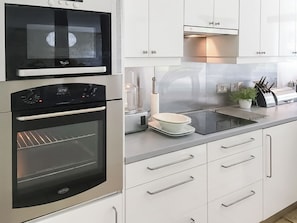 A modern kitchen with stainless steel appliances, including a microwave, oven, and toaster. There are white cabinets with silver handles, a glass-top island, and a potted plant on the counter. - 4 bedroom accommodation in Shilton (Shilton)